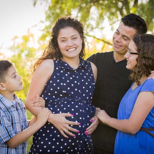 A pregnant mother in a polka dot dress stands with her husband and two children outside in a park.