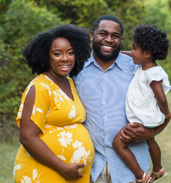 A pregnant mother, her daughter, and her husband stand together in a serene park.
