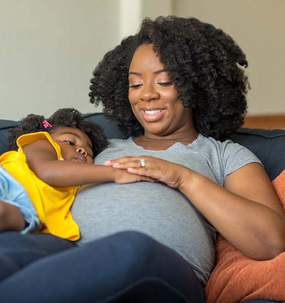A pregnant mother and her daughter sit together on a family sofa.